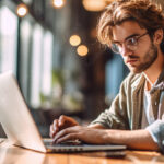 Junger Mann arbeitet am Laptop, Junge Freiberufler oder Student mit Computer im Café am Tisch und schaut in die Kamera.